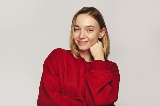 Blonde caucasian young girl smiles positively, wears red sweatshirt holds chin with hand, isolated over white background