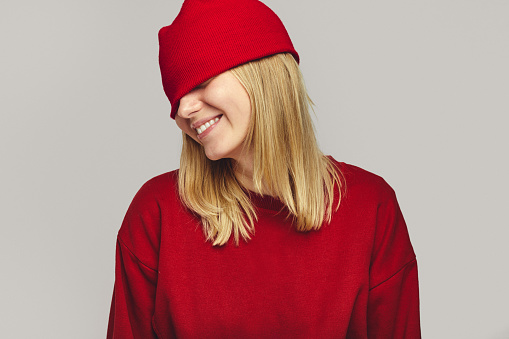 Cheerful hipster female in red outfit, covering eyes with hat, smiling broadly on white background, positive emotions