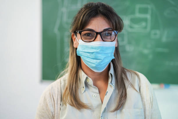 Happy latin teacher smiling on camera while wearing safety masks inside class room - Focus on nose stock photo