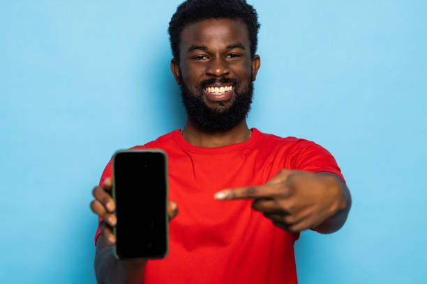 istock Excited young african american man Point index finger on mobile phone with blank empty screen posing isolated on blue background. People lifestyle concept. 1357457232