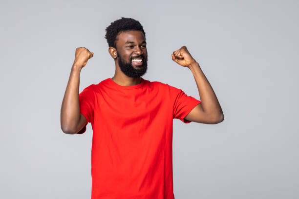 istock Young african american man in blank white t shirt, shouting while his team win, raised both fist in victory gesture, on gray background 1357458105
