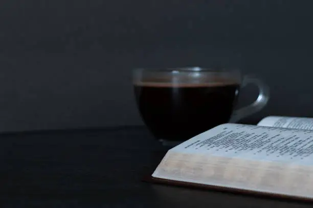 An open Holy Bible Book with a cup of coffee on dark background. Biblical concept of morning prayer to God Jesus Christ, daily studying and reading Scriptures. Christian relationship with the LORD.