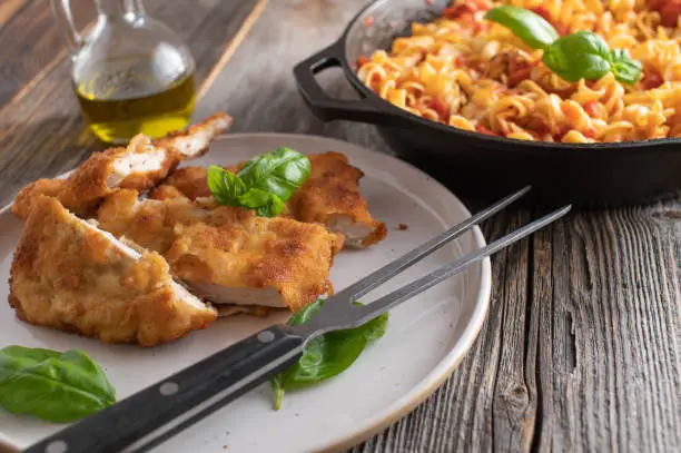 Italian meat dish with pan fried and breaded chicken breast. Served with pasta in a delicious tomato vegetable sauce on wooden table. Closeup
