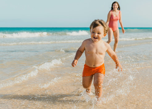 Happy shirtless toddler boy having fun and running on sea waves on sandy beach while playing with mom during summer day