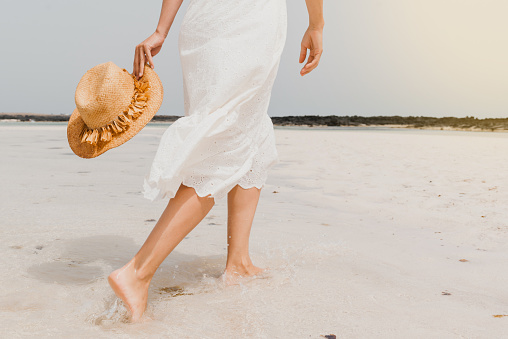 Back view of crop anonymous barefooted female in white dress and with straw hat in hand strolling on wet sandy beach in summer day