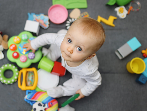 Contented, cute 1 year old baby girl smiling and looking up at the camera surrounded by colourful toys and blocks