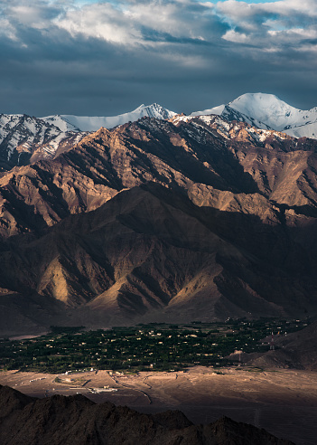 A village in Ladakh at sunrise touched by the morning light and surrounded by Huge snowy Mountains that are light by the morning sun with dark clouds