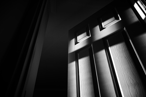 Monochrome image depicting a door slightly ajar, covered with shadows and leading to a shadowy dark room beyond.