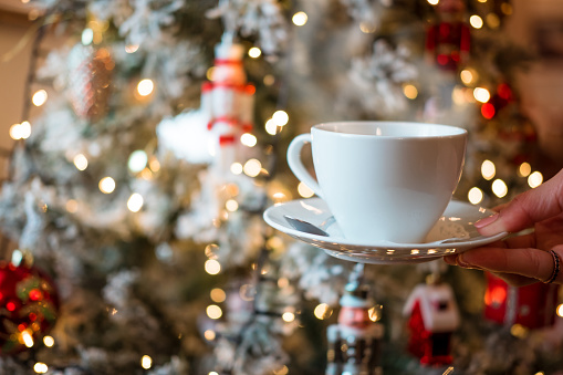 Woman holding a white cup of hot coffee with the decorated Christmas tree in the background. Christmas tree lights create a bokeh effect to celebrate the holy festivities.