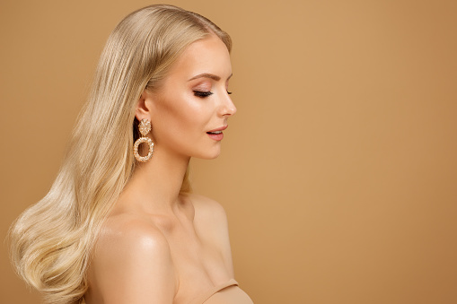 Beauty Blonde Hair Model Face over Beige. Elegant Woman Portrait with Gold Earring. Fashion Girl with Natural Make up and Glossy Skin Profile View