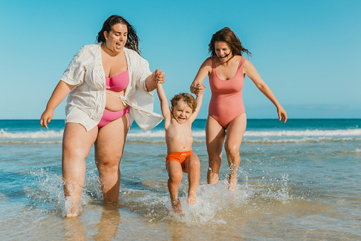 Full body cheerful women in swimwear holding hands and swinging excited boy while standing in waving sea water on sunny day on resort