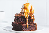Stack of brownie squares with scoop of ice cream and caramel, white background.