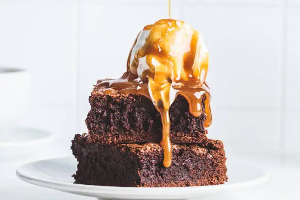 Photo of Stack of brownie squares with scoop of ice cream and caramel, white background.