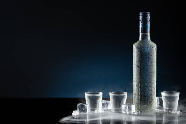 ice-cold bottle of vodka with shot glasses on dark blue background with copy space. - shot glass imagens e fotografias de stock