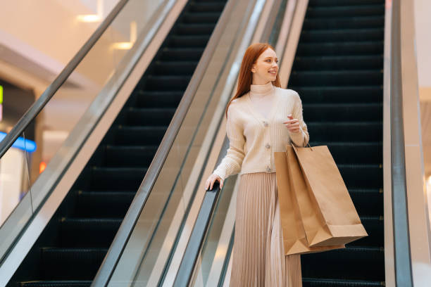 Low-angle view of smiling young woman holding on escalator handrail and riding escalator going up in shopping mall, looking away. Low-angle view of smiling young woman holding on escalator handrail and riding escalator going up in shopping mall, looking away, paper bags with purchases in hands, blurred background. mall walker stock pictures, royalty-free photos & images