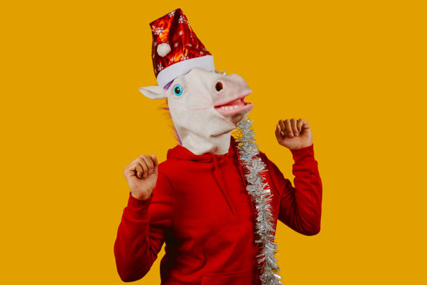 a young man is dancing, wearing a unicorn mask, a santa hat and a strip of silver tinsel on an orangish yellow background, with some blank space around him