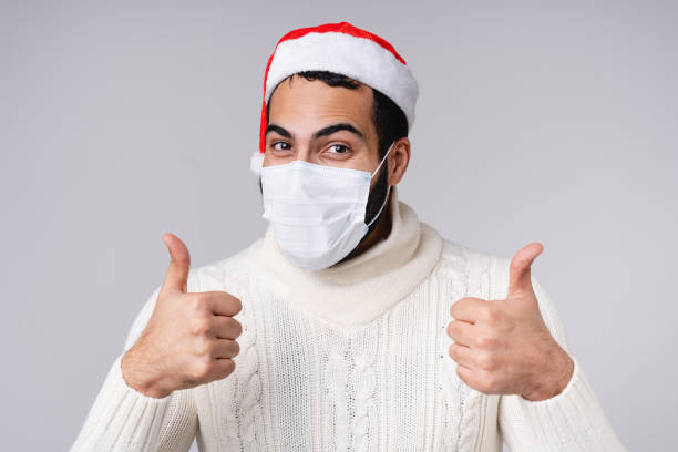 un homme arabe joyeux en masque de protection contre le coronavirus covid19 montrant le pouce levé à noël nouvel an chapeau rouge isolé sur fond gris - coughing illness men latin american and hispanic ethnicity photos et images de collection