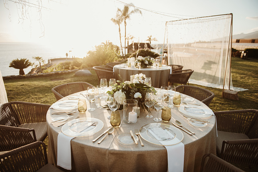 Round tables with elegant dishware and decorations located on coast near sea before wedding banquet at sundown
