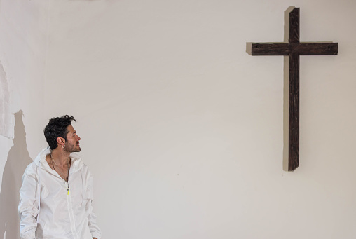 Young latin man dressed in white looking towards a wooden cross in white background