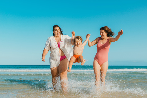 Full body happy women smiling and swinging delighted boy over waving sea water against cloudless blue sky on resort