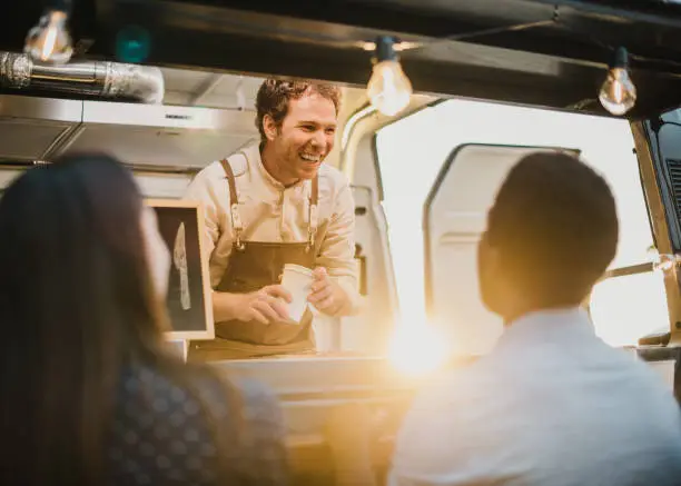 Delighted man with takeaway cups smiling and talking with diverse couple while working in food truck in evening on street