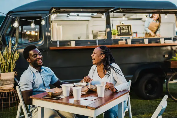 Delighted African American man and woman sitting at table with takeaway food and laughing at joke during dinner near food truck in park in evening