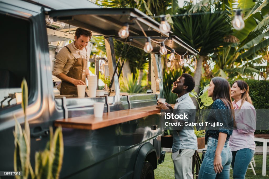Multiracial friends making order to seller in food truck Multiethnic friends standing in queue and ordering takeaway dishes from man in apron working in food truck in summer in park Food Truck Stock Photo