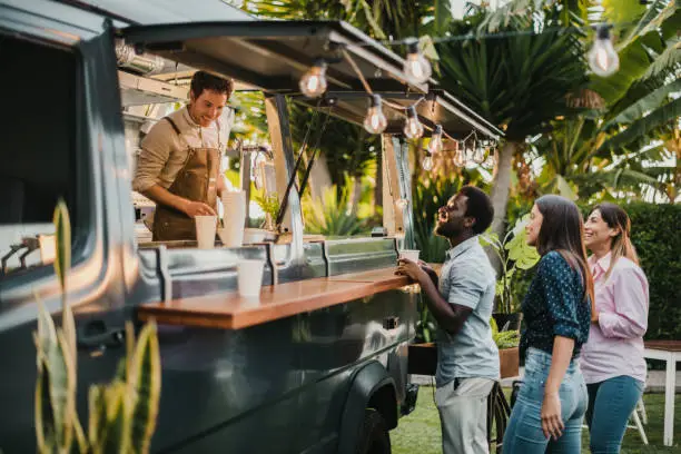 Multiethnic friends standing in queue and ordering takeaway dishes from man in apron working in food truck in summer in park