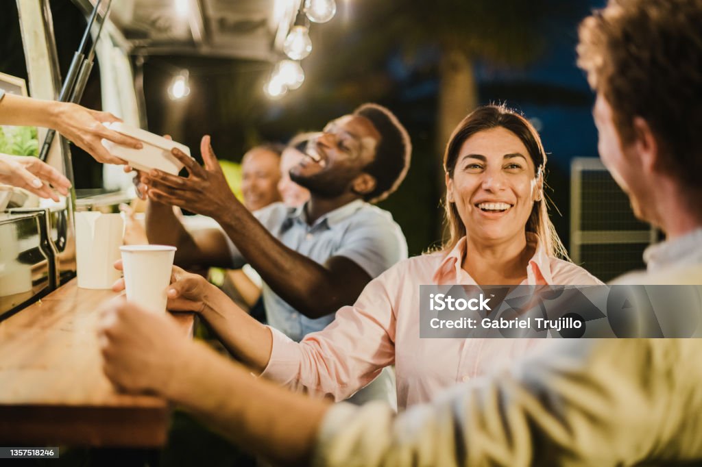 Hispanic woman speaking with friend outside food truck. Happy Hispanic female with takeaway drink smiling and talking with male friend while resting outside food truck in evening Food Truck Stock Photo