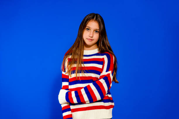 adorable eleven years old girl wearing a red and blue striped sweater on blue studio background, pretty brown-haired fringe hairstyle european appearance child pose indoor smiling look at camera, generation z concept - 10 11 years child human face female imagens e fotografias de stock