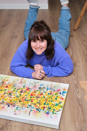 Young woman laughing, looking at camera and painting on canvas with oil paints at home. Artist drawing laying on the floor.