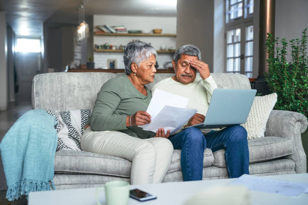 shot of a senior couple looking stressed while using a laptop and going through paperwork on the sofa at home - latin american and hispanic ethnicity senior adult mature adult couple imagens e fotografias de stock