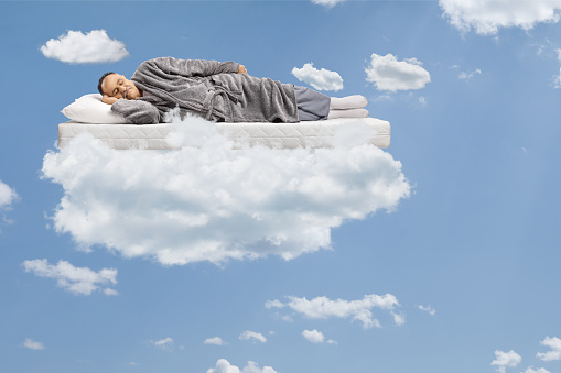 Mature man sleeping on a matress and floating in a blue sky with clouds