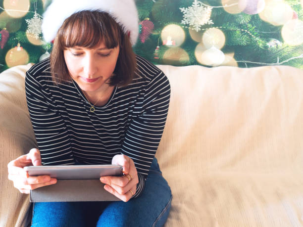 istock Young woman wearing santa cap using tablet on home couch 1357523264