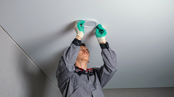 Professional short-haired electrician in work gloves and grey jacket installs light on ceiling standing on ladder during apartment renovation.