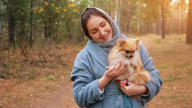 Photo of Young woman holding pomeranian mini spitz in arms while walking in autumn park