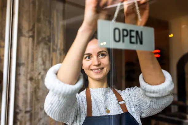 Woman coffee shop owner putting an open sign on the front door. Female barista owner hanging an open sign at a cafe.