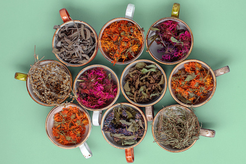 Assortment of dried relaxing tea herbs in colourful cups on mint green background. Calendula, mint, anise hyssop, monarda didyma, wormwood, sage leaves.
