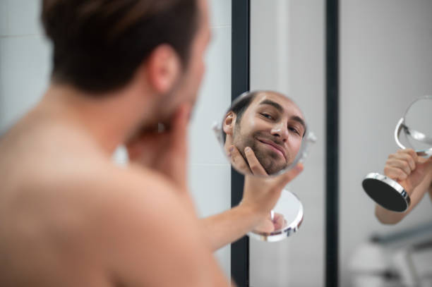 istock Young man examining his face while watching into the round mirror 1357526135