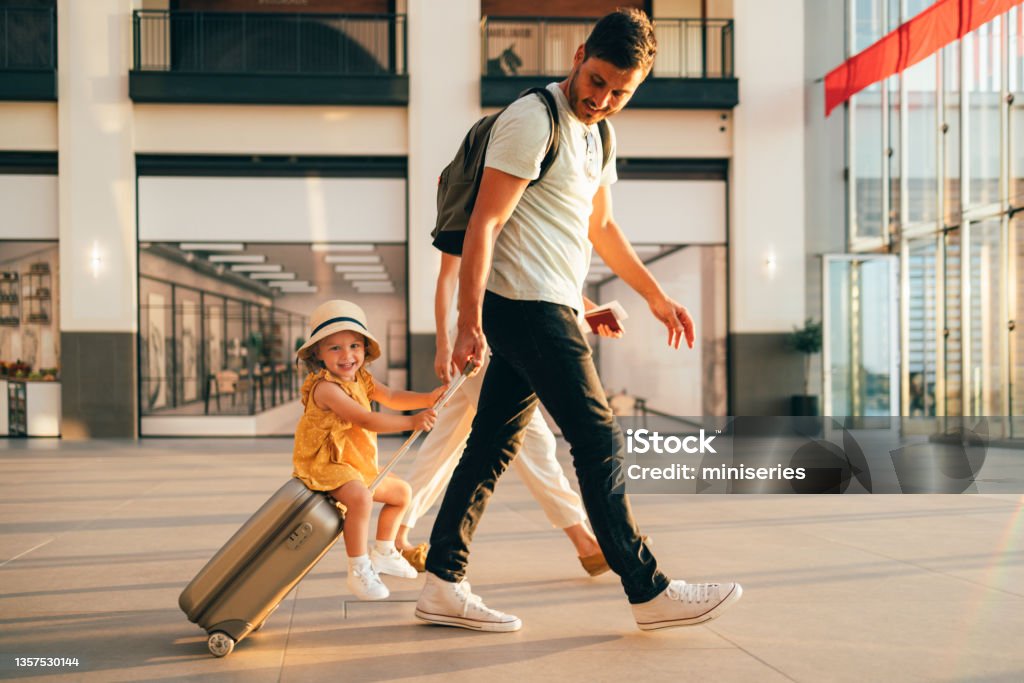 Young Family Having Fun Traveling Together Cheerful husband and his anonymous wife walking with their little girl sitting on luggage at the airport. Travel Stock Photo