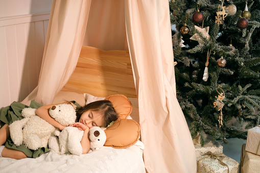 A cute little girl fell asleep on Christmas Eve in her crib waiting to meet Santa. A child sleeps in an embrace with teddy bears. xmas night. Celebrating the New Year. Cozy soft baby bed