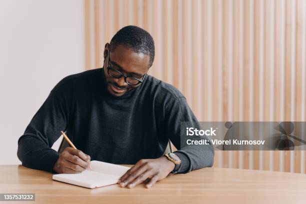 Focused Young African Man In Glasses Holding Pen Taking Notes In Agenda While Sitting At Desk Stock Photo - Download Image Now