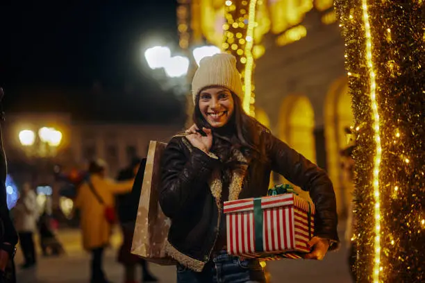 Smiling young adult woman walking through the city streets at Christmas, going from shopping and carrying shopping bags and wrapped Christmas box