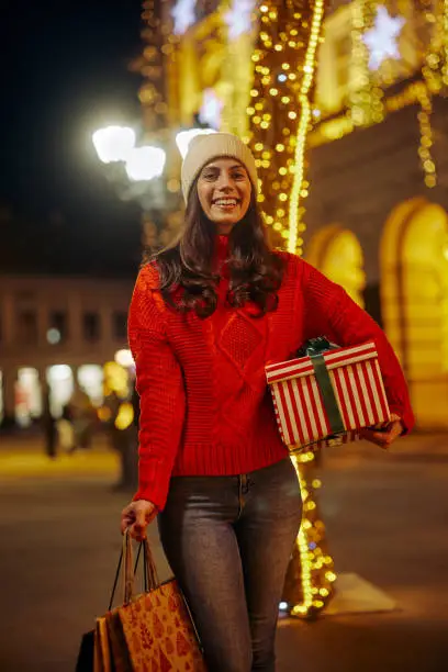 Street portrait of smiling beautiful young woman with wrapped christmas box and shopping bags on the festive Christmas fair.