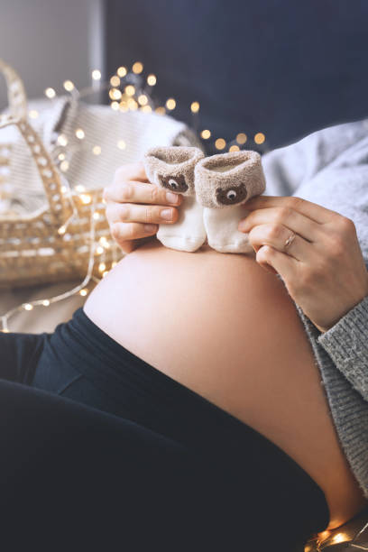 임신 한 여성은 그녀의 배 근처에 작은 아기 양말을 들고. 임신 중 신생아를위한 초음파 이미지와 물건으로 고리버들 바구니를 준비하는 임산부. - human pregnancy baby shower image color image 뉴스 사진 이미지