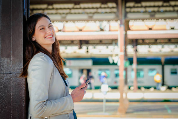 istock Smiling business woman using smartphone while waiting in a train station - Attractive young student girl using cell-phone while standing on the railway station platform 1357543051