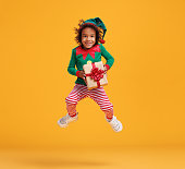 Happy african american kid boy Christmas elf costume with wrapped Xmas gift in hands jumping in air