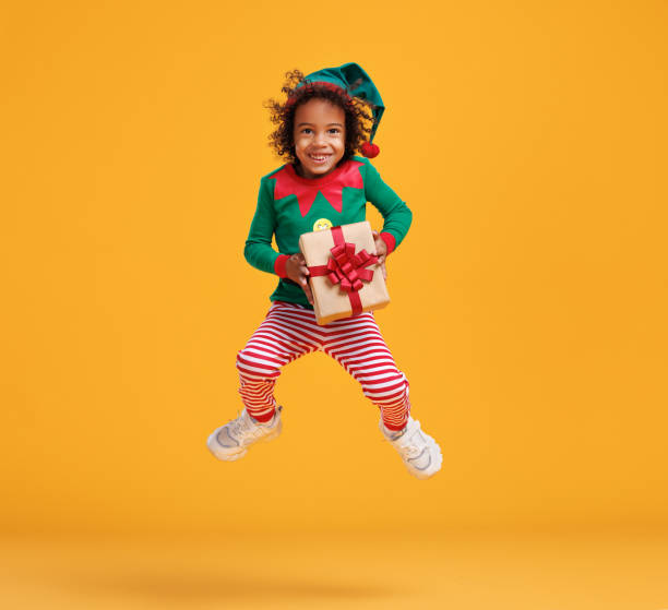 Happy african american kid boy Christmas elf costume with wrapped Xmas gift in hands jumping in air Little santa claus helper. Photo in motion of happy african american kid boy Christmas elf costume with wrapped Xmas gift in hands jumping up in air isolated over orange studio wall santa claus elf assistance christmas stock pictures, royalty-free photos & images