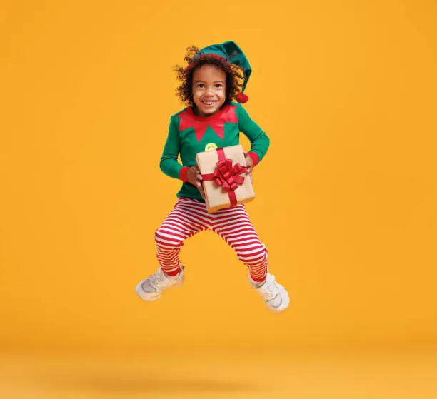 Little santa claus helper. Photo in motion of happy african american kid boy Christmas elf costume with wrapped Xmas gift in hands jumping up in air isolated over orange studio wall