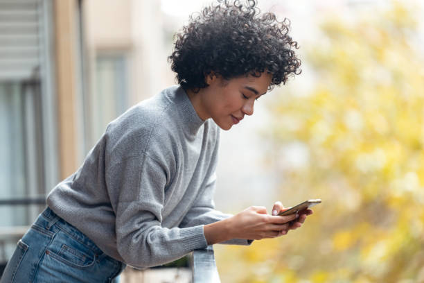 istock Cheerful woman using her smartphone while sending messages standing in the balcony at home. 1357546496